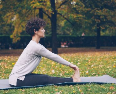 Slim young lady is doing sequence of yoga asanas sitting on mat in park alone on warm autumn day with beautiful grass and trees around her. Recreation and sport concept.