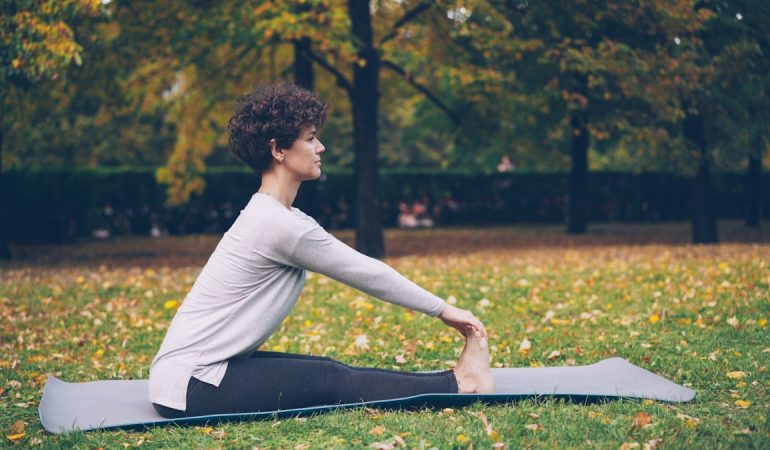 Slim young lady is doing sequence of yoga asanas sitting on mat in park alone on warm autumn day with beautiful grass and trees around her. Recreation and sport concept.