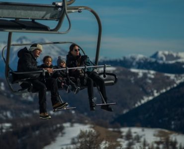 Family having fun on a ski lift with snow-covered mountains in the background during a sunny winter day.