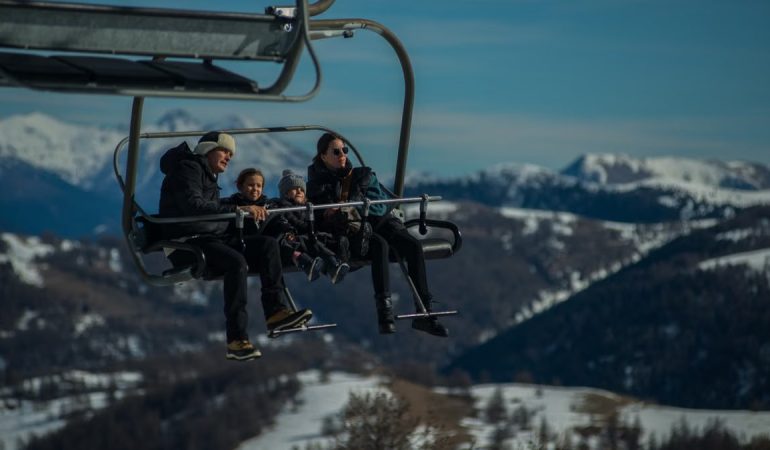 Family having fun on a ski lift with snow-covered mountains in the background during a sunny winter day.